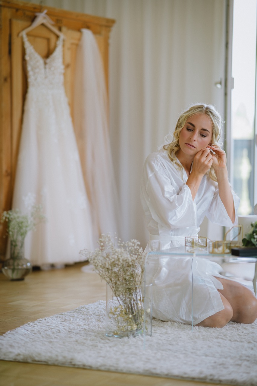 Braut legt sich Ohrringe an — Brautkleid hängt im Hintergrund