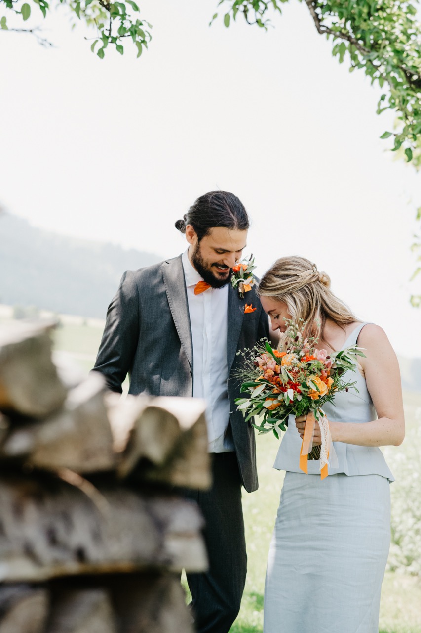 Bräutigam und Braut lachen gemeinsam mit Wildblumenstrauss im Grünen