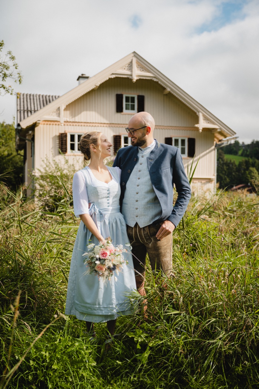 Brautpaar in Dirndl und Lederhose steht in einer hohen Wiese vor einem Bauernhaus